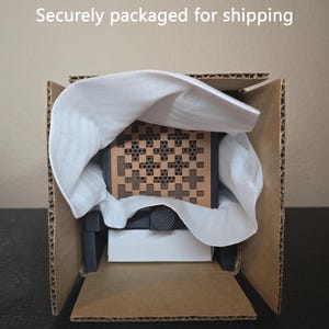 May include: A square object with a brown, perforated geometric pattern featuring cross shapes, partially wrapped in white protective foam inside a brown cardboard box. The text "Securely packaged for shipping" is visible at the top.