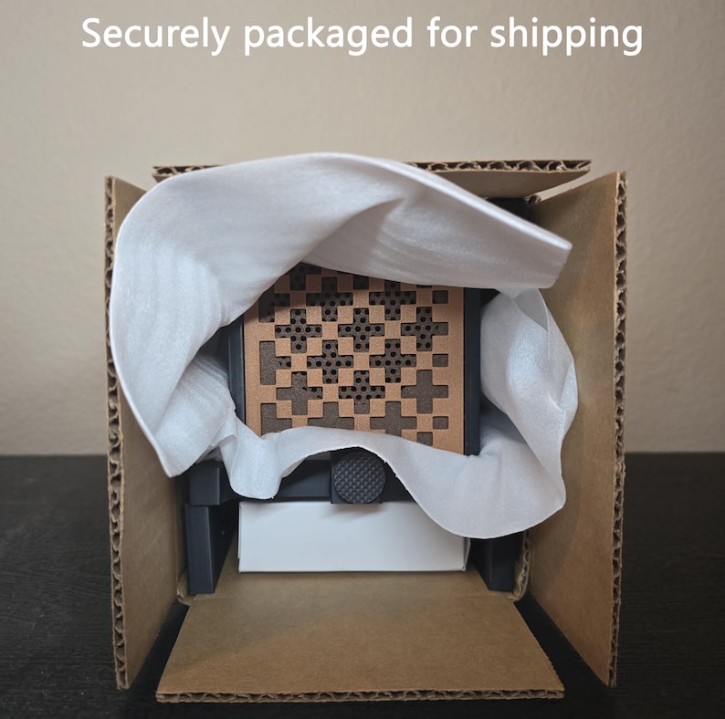 May include: A square object with a brown, perforated geometric pattern featuring cross shapes, partially wrapped in white protective foam inside a brown cardboard box. The text "Securely packaged for shipping" is visible at the top.