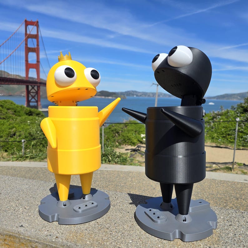 May include: Two stylized character figures standing on gray circular bases. One figure is bright yellow with a small crown, and the other is matte black. Both have large white eyes with black pupils, cylindrical bodies, and pointed arms.