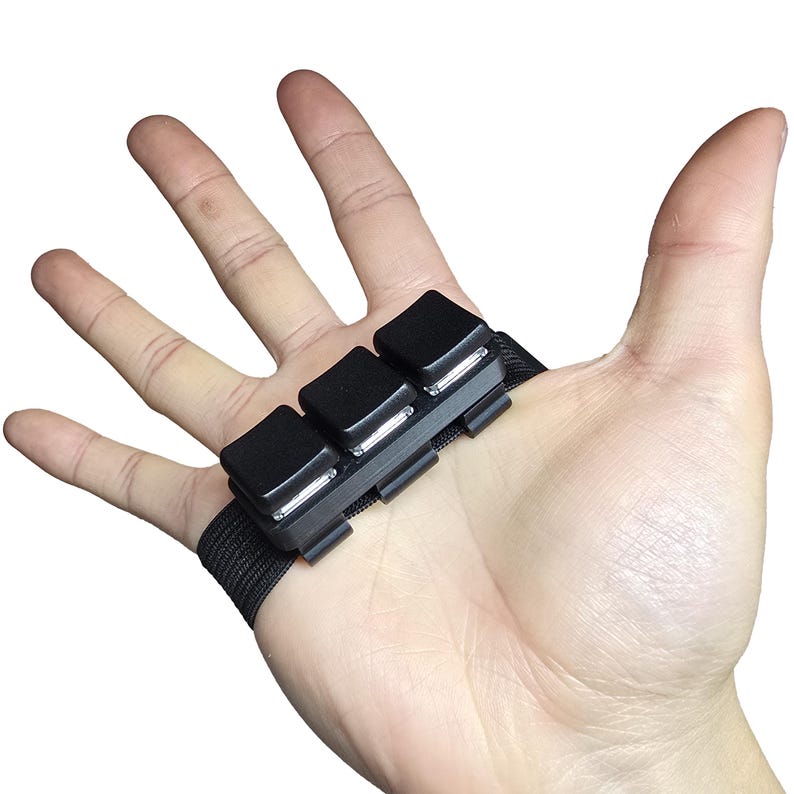 May include: A black finger-mounted device with three square buttons and a black strap. The device is designed to be worn on the finger and is likely used for gaming or other digital interactions.