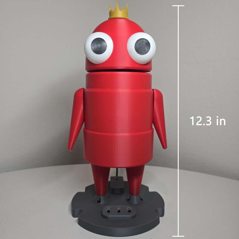 May include: A red robot-like figure with large white eyes and a small gold crown on its head. The figure stands on a dark gray circular base and measures 12.3 inches in height. It features a cylindrical body, two arms, and two legs.