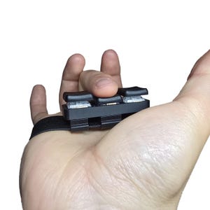May include: A black, wearable keyboard with three keys, held in a hand. The keyboard is attached to a black wrist strap. The keys are black and have a clear, rectangular component visible beneath them.