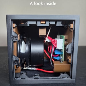 May include: A black square electronic housing with the text A look inside printed at the top. The interior reveals a black speaker, brown structural supports, a green circuit board, and red wiring connected to white plastic components and screws.