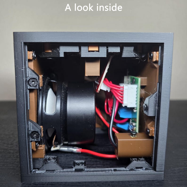 May include: A black square electronic housing with the text A look inside printed at the top. The interior reveals a black speaker, brown structural supports, a green circuit board, and red wiring connected to white plastic components and screws.