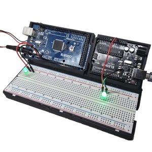 以下が含まれることがあります： 黒と青の回路基板に接続された、赤と緑のLEDが2つ付いたブレッドボード。回路基板には「ELEGOO UNO R3」と印刷されています。