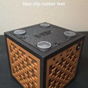May include: A black and brown cube-shaped device featuring a textured grid pattern on its sides. The top surface is black, labeled with the text 1-TEK, and includes four circular non-slip rubber feet secured by small screws at each corner.