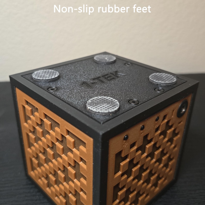 May include: A black and brown cube-shaped device featuring a textured grid pattern on its sides. The top surface is black, labeled with the text 1-TEK, and includes four circular non-slip rubber feet secured by small screws at each corner.