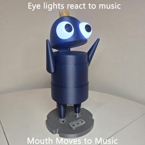 May include: A dark blue robot figure with a small gold crown on its head, large glowing white eyes, and raised arms. The figure stands on a gray circular base. Text on the image reads: Eye lights react to music and Mouth Moves to Music.