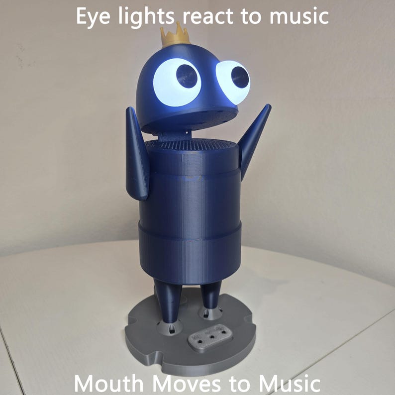 May include: A dark blue robot figure with a small gold crown on its head, large glowing white eyes, and raised arms. The figure stands on a gray circular base. Text on the image reads: Eye lights react to music and Mouth Moves to Music.