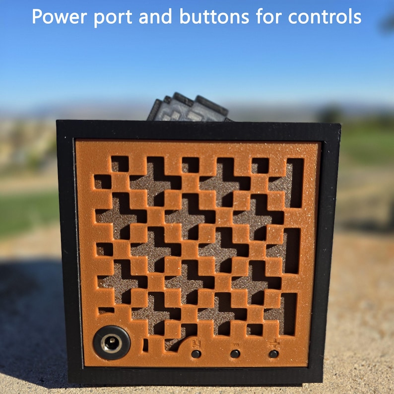 May include: A square electronic device with a black frame and a copper-colored front panel featuring a geometric cross-shaped cutout pattern. The panel includes a circular power port and three small control buttons. Text reads: Power port and buttons for controls.