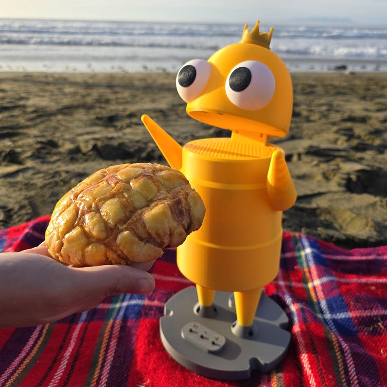 May include: A yellow, cylindrical, robot-like figure with large white eyes, a small crown on top, and a gray circular base, positioned next to a golden-brown, textured, round pastry with a grid-like pattern on its surface.