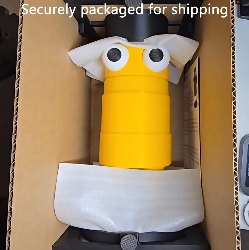 May include: A yellow cylindrical object with two large white and black googly eyes, positioned inside a cardboard box with protective white foam padding. The text "Securely packaged for shipping" is visible at the top of the image.