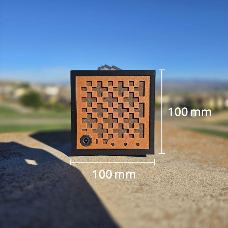 May include: A square black and brown box with a repeating cross pattern on the front panel. The device features a circular port in the lower left corner and three small circular dots. It measures 100 mm in height and 100 mm in width.