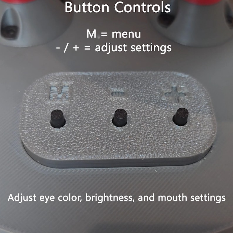 May include: A gray control panel with three black buttons and embossed symbols M, -, and +. Text reads: Button Controls, M = menu, - / + = adjust settings, Adjust eye color, brightness, and mouth settings. The surface has a textured finish.