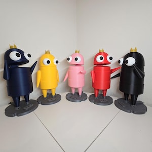 May include: Five standing robot-like figures in dark blue, yellow, pink, red, and black. Each figure has a cylindrical body, two large white eyes with black pupils, and a small gold crown on top. They are mounted on individual gray bases.