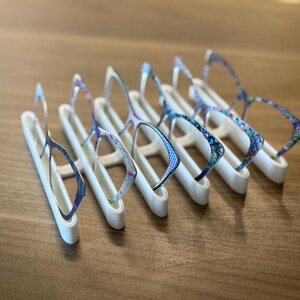 Puede incluir: Una colecci&oacute;n de gafas con monturas estampadas, expuestas sobre soportes blancos. Las monturas presentan dise&ntilde;os azules, morados y blancos. Los soportes est&aacute;n dispuestos sobre una superficie de madera.