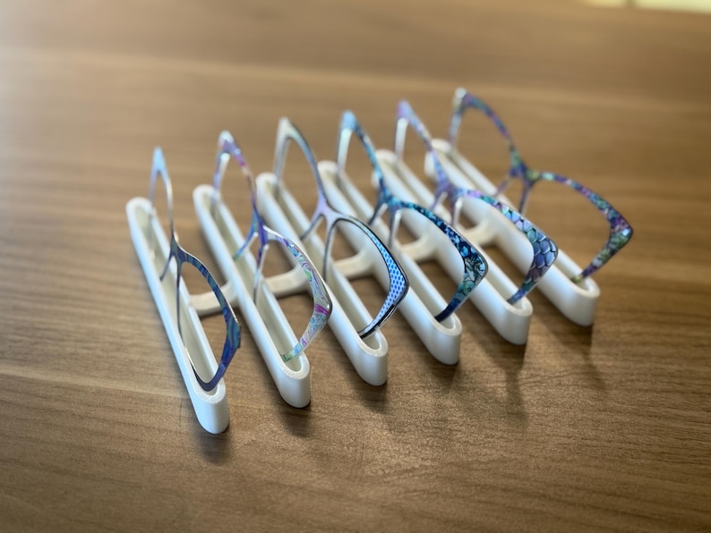 Puede incluir: Una colecci&oacute;n de gafas con monturas estampadas, expuestas sobre soportes blancos. Las monturas presentan dise&ntilde;os azules, morados y blancos. Los soportes est&aacute;n dispuestos sobre una superficie de madera.