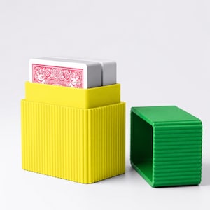 May include: A bright yellow rectangular storage box with vertical ribbed sides containing a deck of playing cards with a red back design. A separate green rectangular lid with matching vertical ribbed sides sits to the right.