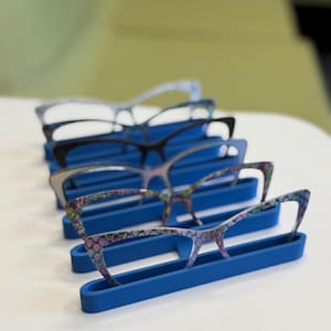 Puede incluir: Una colecci&oacute;n de gafas con varios dise&ntilde;os de montura, incluyendo estilos florales y estampados, exhibidas en soportes azules. Las gafas est&aacute;n dispuestas en fila, mostrando diferentes formas y colores.