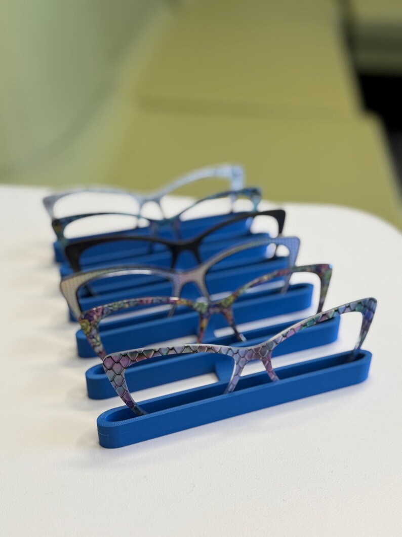 Puede incluir: Una colecci&oacute;n de gafas con varios dise&ntilde;os de montura, incluyendo estilos florales y estampados, exhibidas en soportes azules. Las gafas est&aacute;n dispuestas en fila, mostrando diferentes formas y colores.