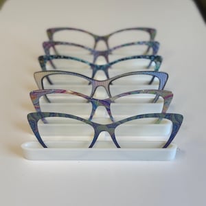 Puede incluir: Una exhibici&oacute;n de cinco gafas de ojo de gato con patrones coloridos y arremolinados. Las monturas est&aacute;n dispuestas sobre un soporte rectangular blanco. Las gafas tienen un estilo retro con un toque moderno, mostrando una variedad de colores vibrantes.