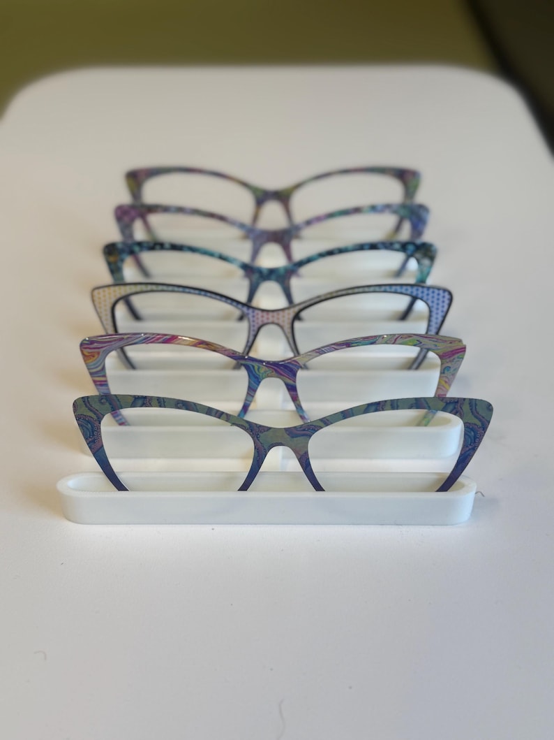Puede incluir: Una exhibici&oacute;n de cinco gafas de ojo de gato con patrones coloridos y arremolinados. Las monturas est&aacute;n dispuestas sobre un soporte rectangular blanco. Las gafas tienen un estilo retro con un toque moderno, mostrando una variedad de colores vibrantes.