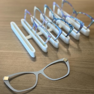 Puede incluir: Una exhibici&oacute;n de gafas y un soporte blanco para gafas. Las gafas tienen una montura gris claro con detalles dorados. Varios pares de gafas con monturas estampadas de colores se exhiben en el soporte.