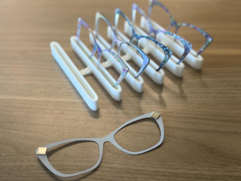 Puede incluir: Una exhibici&oacute;n de gafas y un soporte blanco para gafas. Las gafas tienen una montura gris claro con detalles dorados. Varios pares de gafas con monturas estampadas de colores se exhiben en el soporte.