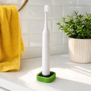 May include: A white electric toothbrush standing upright in a square, lime green silicone base with a black ridged interior. The toothbrush features a slim, cylindrical handle with a single circular power button and a standard brush head.