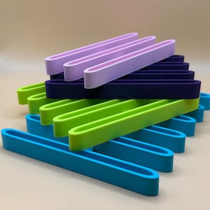 Puede incluir: Una pila de soportes rectangulares de colores, abiertos en los extremos, en tonos de azul, verde, morado y marino. Los soportes est&aacute;n dispuestos en un patr&oacute;n en capas, mostrando sus colores vibrantes y su dise&ntilde;o sencillo. Los soportes son de un material liso.