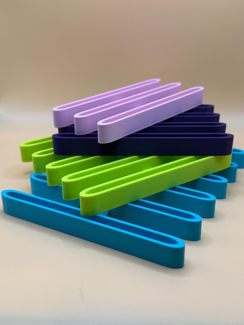 Puede incluir: Una pila de soportes rectangulares de colores, abiertos en los extremos, en tonos de azul, verde, morado y marino. Los soportes est&aacute;n dispuestos en un patr&oacute;n en capas, mostrando sus colores vibrantes y su dise&ntilde;o sencillo. Los soportes son de un material liso.