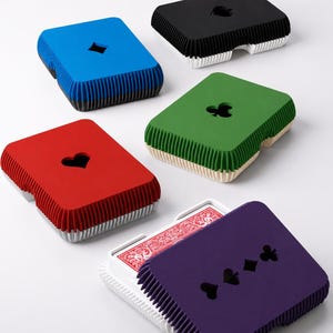 May include: Five rectangular playing card cases with ribbed sides and cut-out suit symbols on the lids. The cases are blue, black, green, red, and purple. One purple case is open, revealing a deck of playing cards with a red patterned back design.