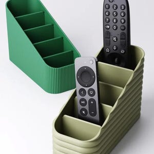 May include: Two remote control organizers in shades of green. One is a bright green with vertical ridges, the other is a muted green. Each has four compartments. Two black remotes are in the muted green organizer.