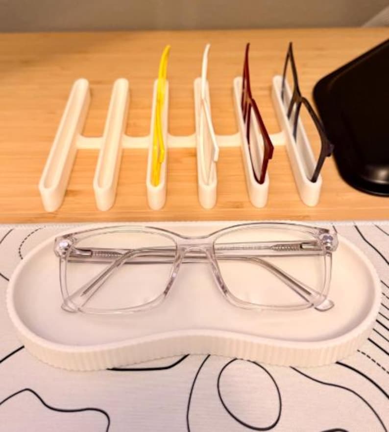 Puede incluir: Un organizador de gafas blanco con ranuras que sostienen varios pares de gafas con monturas amarillas, blancas, rojas y negras. Un par de gafas con montura transparente descansa en una bandeja blanca con bordes ondulados.