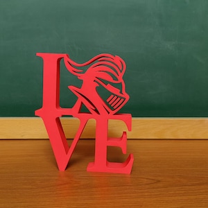 May include: A red decorative sign spelling "LOVE" with a knight's helmet design replacing the "O". The sign is made of a solid material and sits on a wooden surface, with a green chalkboard background.