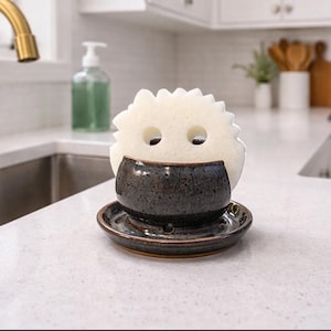 May include: A ceramic sponge holder with a dark blue and brown speckled finish, holding a white sponge with two eye-like holes. The holder sits on a matching saucer, perfect for kitchen organization.