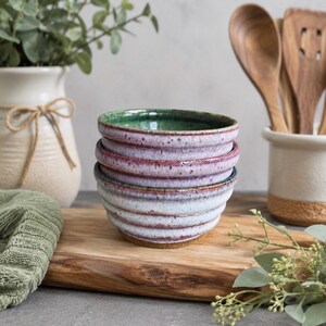 May include: A stack of three ceramic bowls with a ribbed exterior texture. The bowls feature a reactive glaze with a gradient of colors, transitioning from a deep green interior to a purple and white exterior with speckled brown accents.