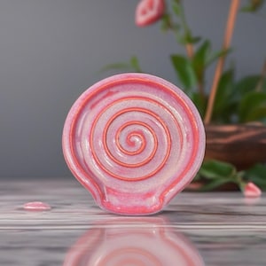May include: A pink ceramic spoon rest with a spiral design in the center. The spoon rest has a small lip for holding utensils and is a light pink color with a darker pink spiral.