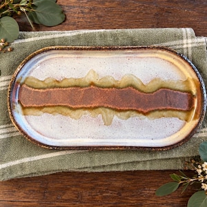 May include: An oval ceramic tray featuring a speckled white glaze with a prominent, horizontal band of deep brown and olive green running through the center. The edges of the tray are finished in a dark, toasted brown tone, creating a distinct border.