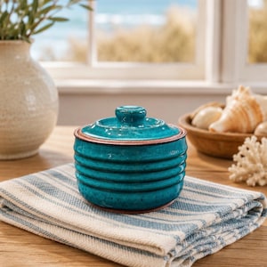 May include: A round, teal-colored ceramic jar with a matching lid, featuring horizontal ribbed detailing along the sides and a smooth, glossy finish. The jar has a visible terracotta-colored rim at the base and the top edge, providing a contrast.