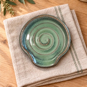 May include: A round ceramic spoon rest featuring a glossy green glaze with a prominent spiral pattern embossed in the center. The edges of the dish are defined by a dark brown rim, creating a contrast against the lighter green interior surface.