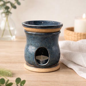 May include: A blue ceramic essential oil warmer with a candle inside. The warmer has a round opening and a separate top piece. The base and the rim of the top are a light tan color. The warmer is on a wooden surface.