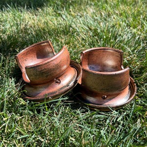 May include: Two handmade ceramic bowls with matching saucers. The bowls and saucers are a reddish-brown color with dark brown and black accents. The bowls have a unique, curved design and sit on the saucers. The bowls are placed on green grass.