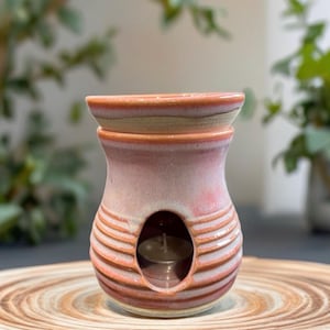May include: A pink and orange ceramic essential oil warmer with a candle inside. The warmer has a round opening for the candle and horizontal stripes. The warmer sits on a wooden, circular tray.
