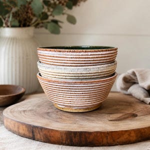 May include: A stack of three ceramic bowls with a ribbed, textured exterior. The bowls feature a white glaze with horizontal brown lines and a speckled finish. The interior of the top bowl is a deep green, while the base of the stack is a tan color.