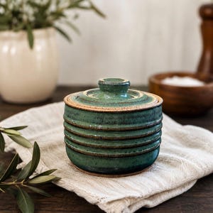 May include: A round, lidded ceramic jar featuring a deep teal and green glaze with visible horizontal ridges along the body. The rim and lid edges show a contrasting earthy brown tone, and the lid is topped with a small, centered knob handle.