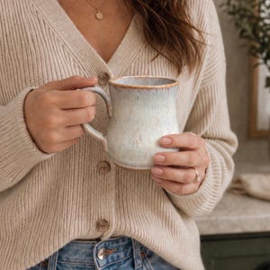 May include: A cream-colored ceramic mug with a speckled glaze and a brown rim is held by a person wearing a beige cardigan and blue jeans. The mug has a curved handle and appears to be filled. A gold necklace is also visible.