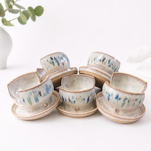 May include: A collection of handmade ceramic bowls and saucers. The bowls are off-white with blue and green speckled patterns. Each bowl sits on a matching saucer, creating a rustic and artistic aesthetic. The set is ideal for serving soups or small dishes.