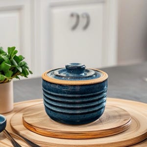 May include: A blue ceramic lidded jar with horizontal ridges, sitting on a wooden serving tray. The jar has a light brown rim and a small knob on the lid. The jar is surrounded by a wooden serving tray and a small plant in a beige pot.