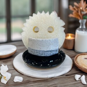 May include: A decorative kitchen sponge with two eye-shaped holes, positioned above a small ceramic cup and a black dish. The cup has blue and white speckled bands. The items are on a white marble-like surface, with a wooden table background.